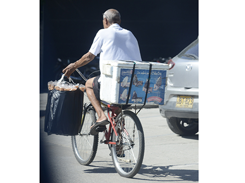 Adulto mayor maneja incómodo su bicicleta con una sola mano por la transitada avenida del Ferrocarril mientras con la izquierda sostiene conos de helado y transporta una ‘neverita’ portátil en su parrilla. 